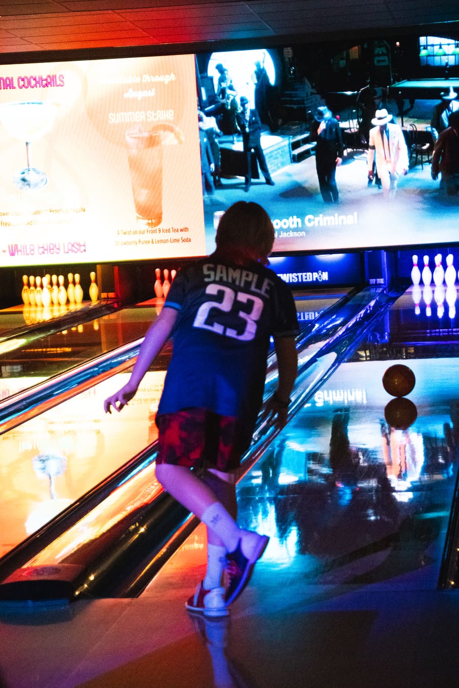 A kid in a #23 jersey releases the ball on a Twisted Pin lane, the LED video wall behind showing cocktail tasting notes